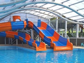 An indoor heated pool with orange and blue waterslides and transparent roof overhead