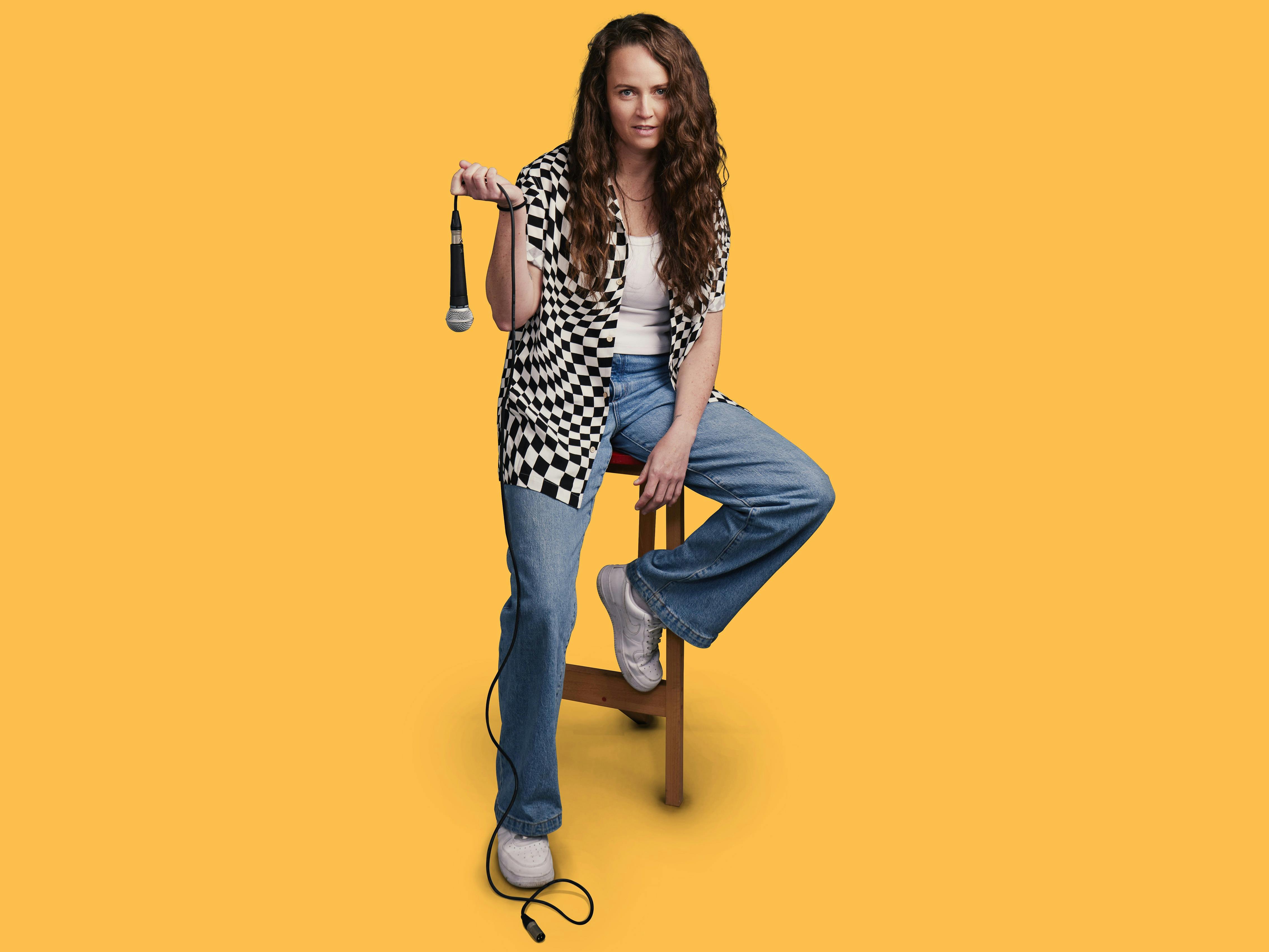 Comedian Zoe Coombs Marr sits on a stool holding a floppy microphone
