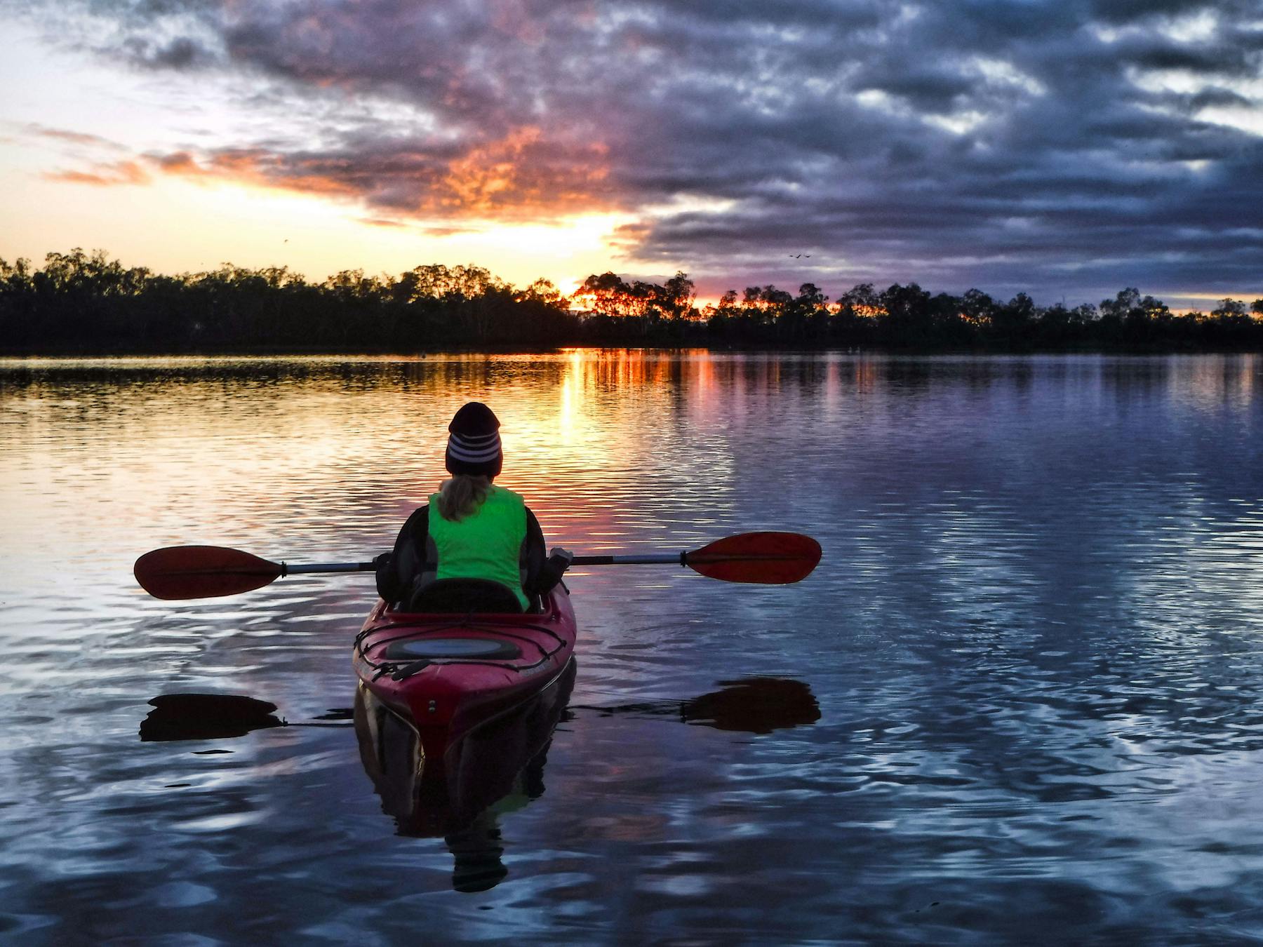 Kayaker in beanie, watching a colourful sunrise from her kayak on a backwater of the Murray River..