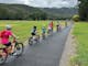A group of children ride bikes in a line along a bitumen path with green grass next to them.