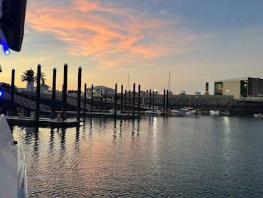 Sunset at marina with boats, jetty, lighthouse, and blue lights on boat reflecting on water.