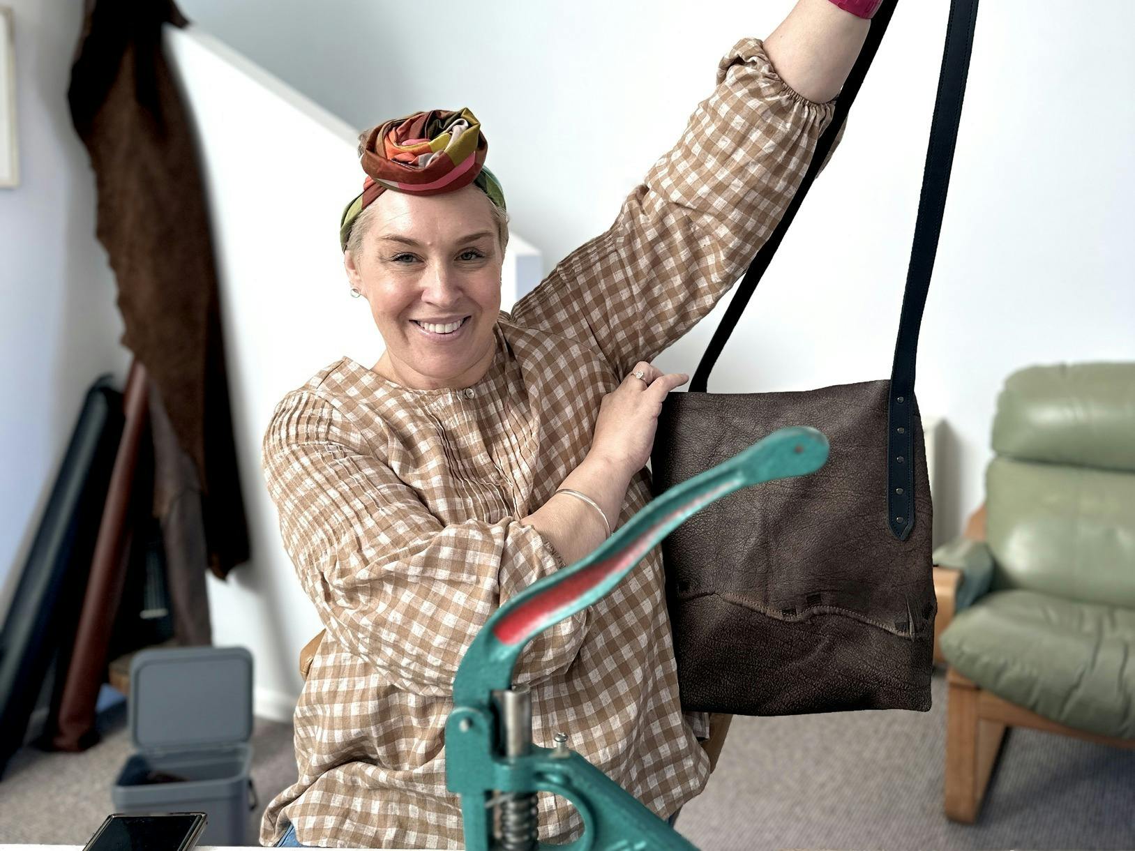 Smiling woman holding up her handmade leather tote bag during a workshop, sitting at a table with le