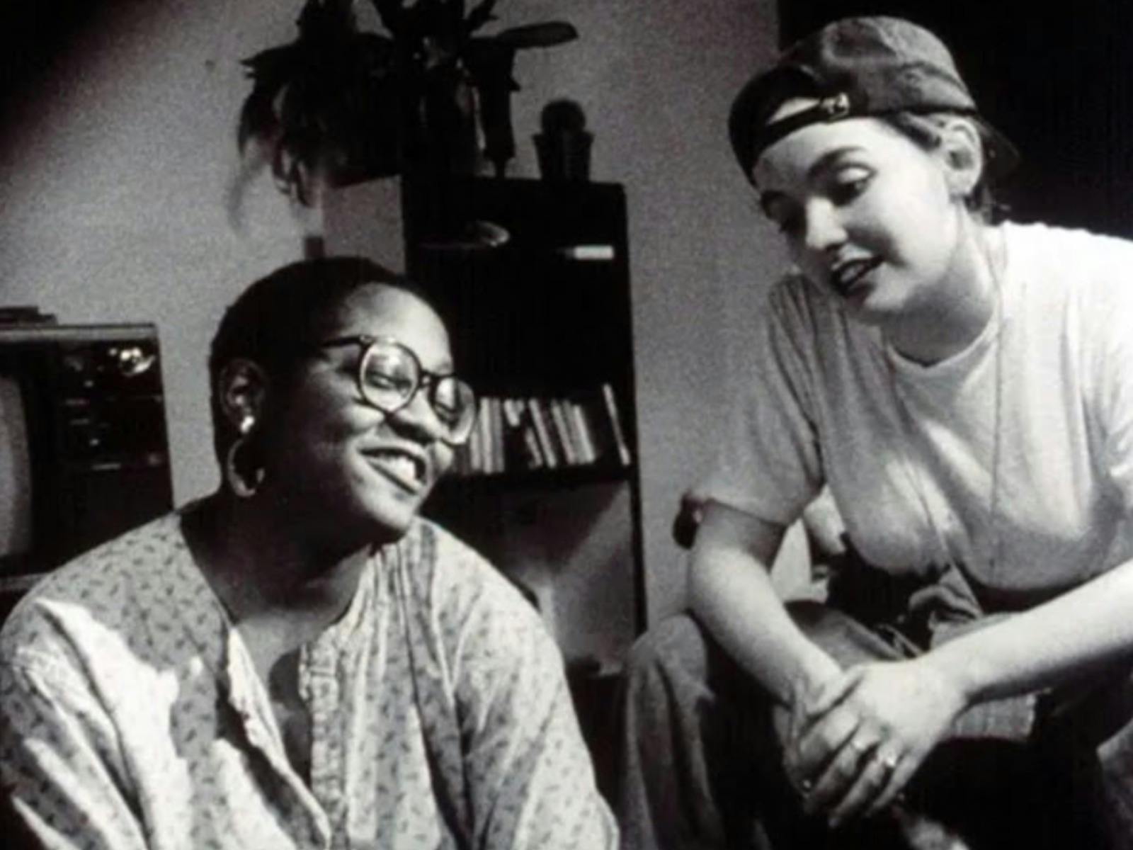 Two people sit together in a room, smiling and talking in a black-and-white scene.