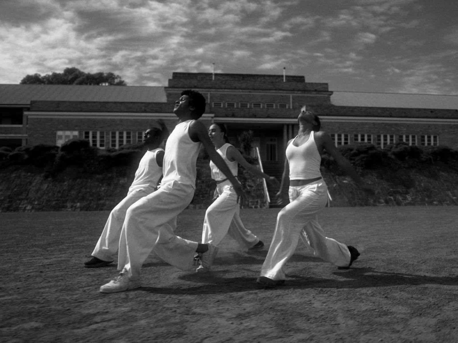 Black and white photo of four people dancing on a field with a building in the background