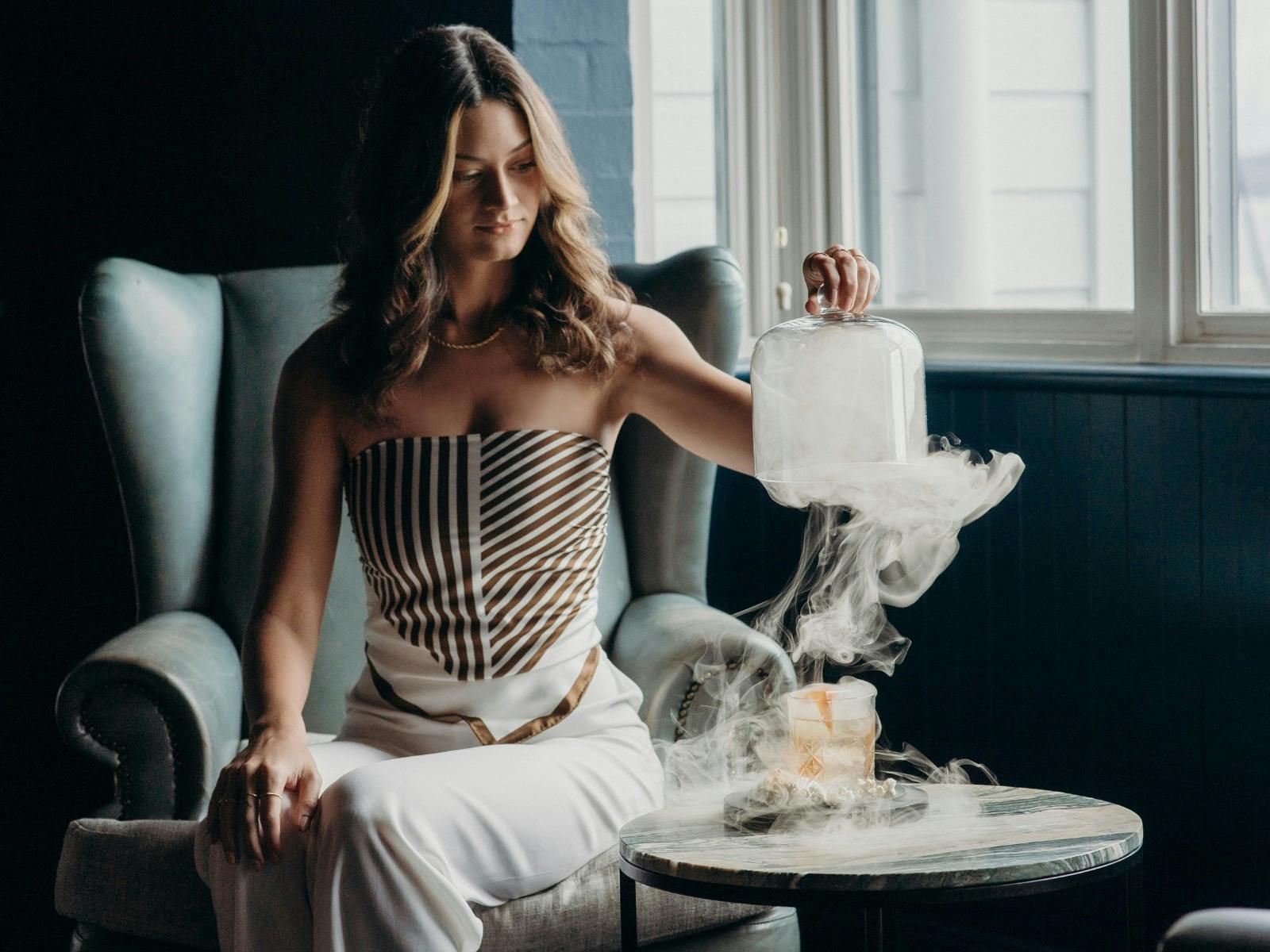 A woman sitting on a lounge chair, lifting a glass closh to reveal a smoky cocktail
