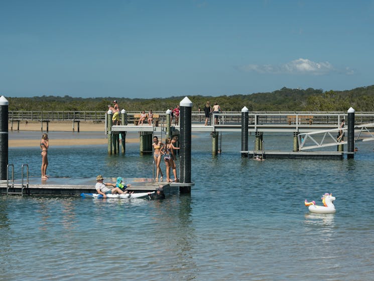 People kayak and swim in an estuary, a group of teenagers stand on a platform above the water.