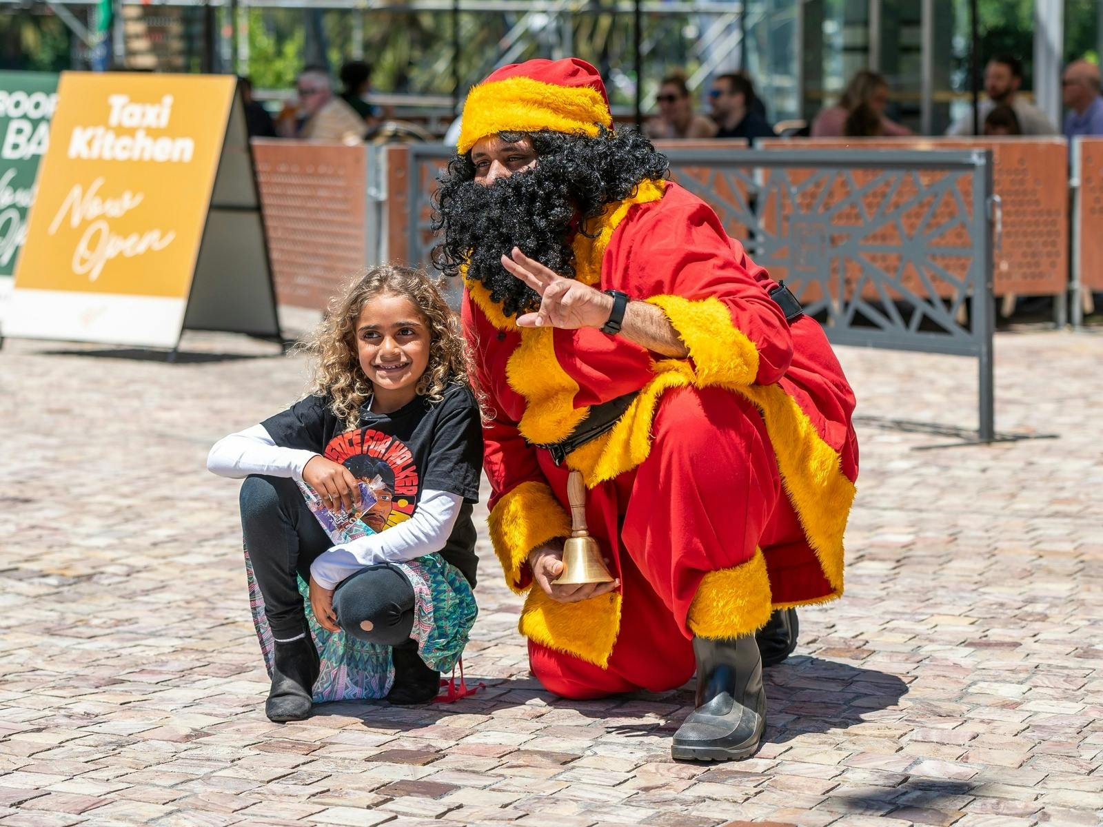 Koorie Klaus is dressed in red and gold and is posing for a photo with a child