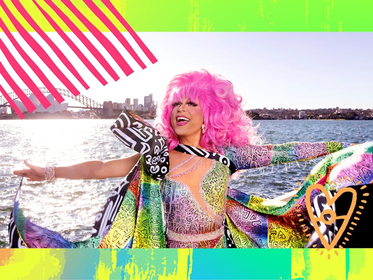 a person in a pink wig and colourful dress with their arms wide smiling with the sea in background