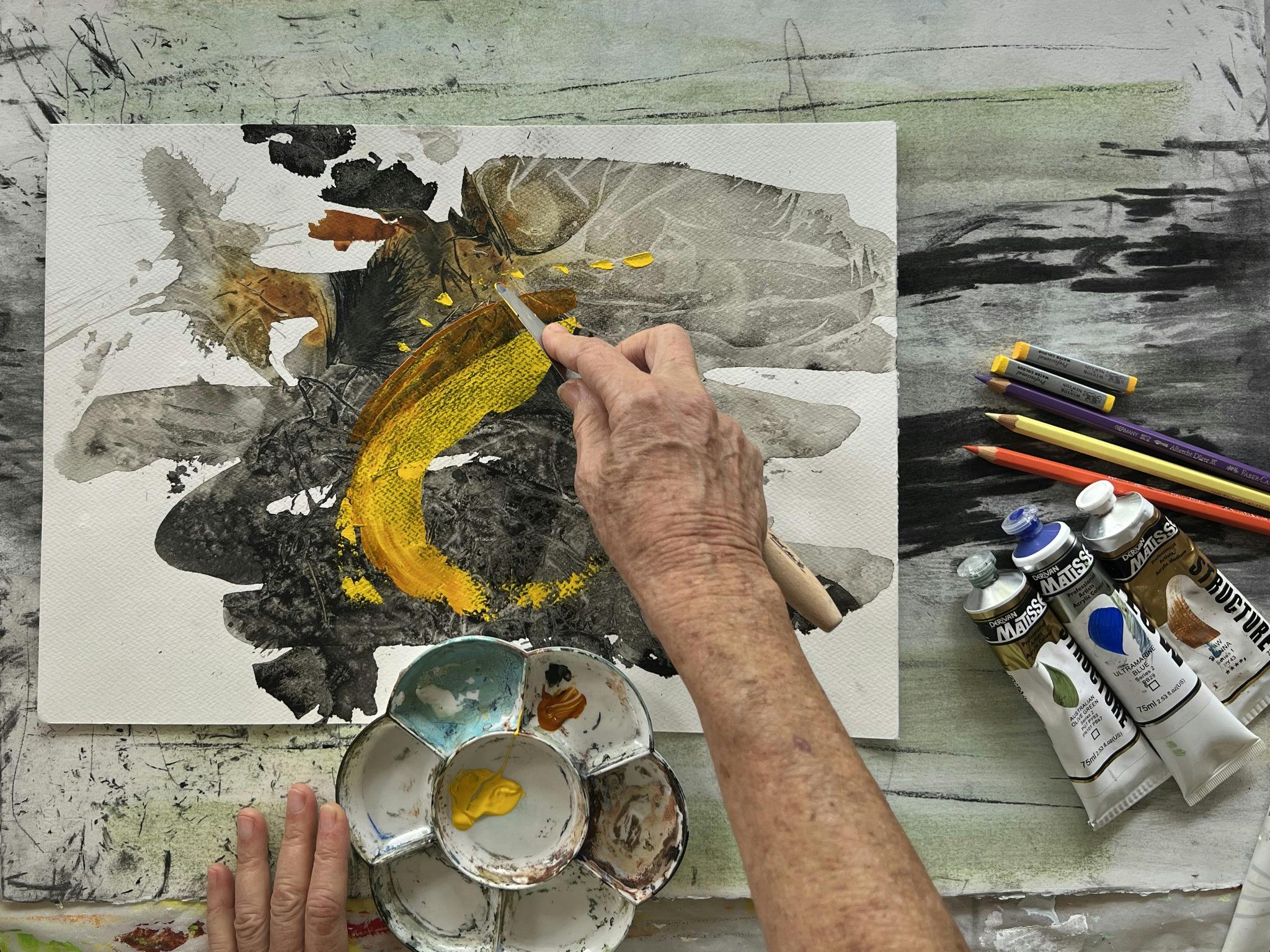 a hand pulls a palette knife across the paper making a mustard yellow mark on a mixed-media artwork