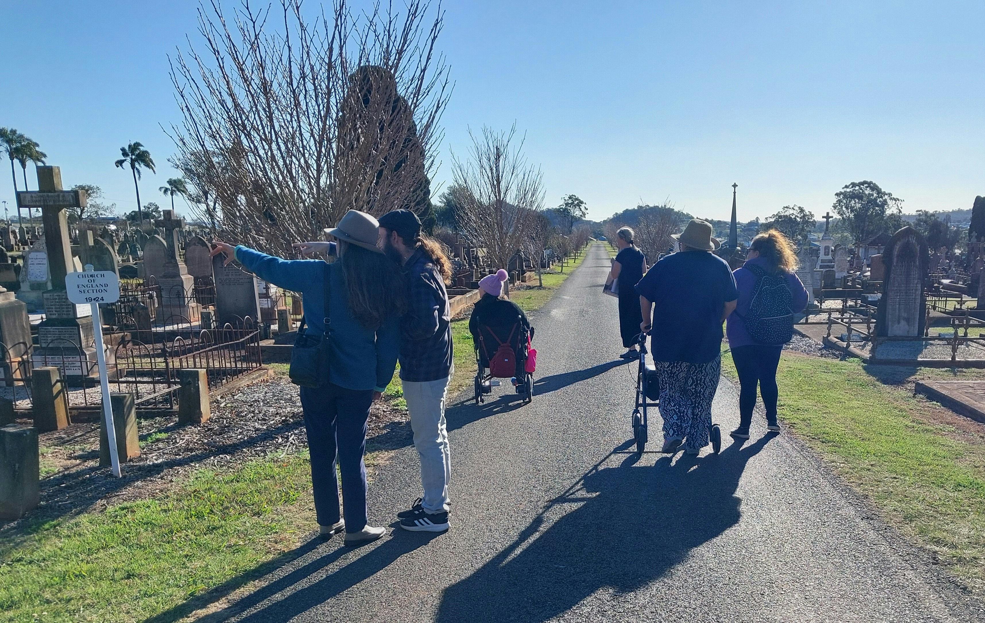 A small group of people, backs to camera, walking and wheeling down a cemetery avenue.