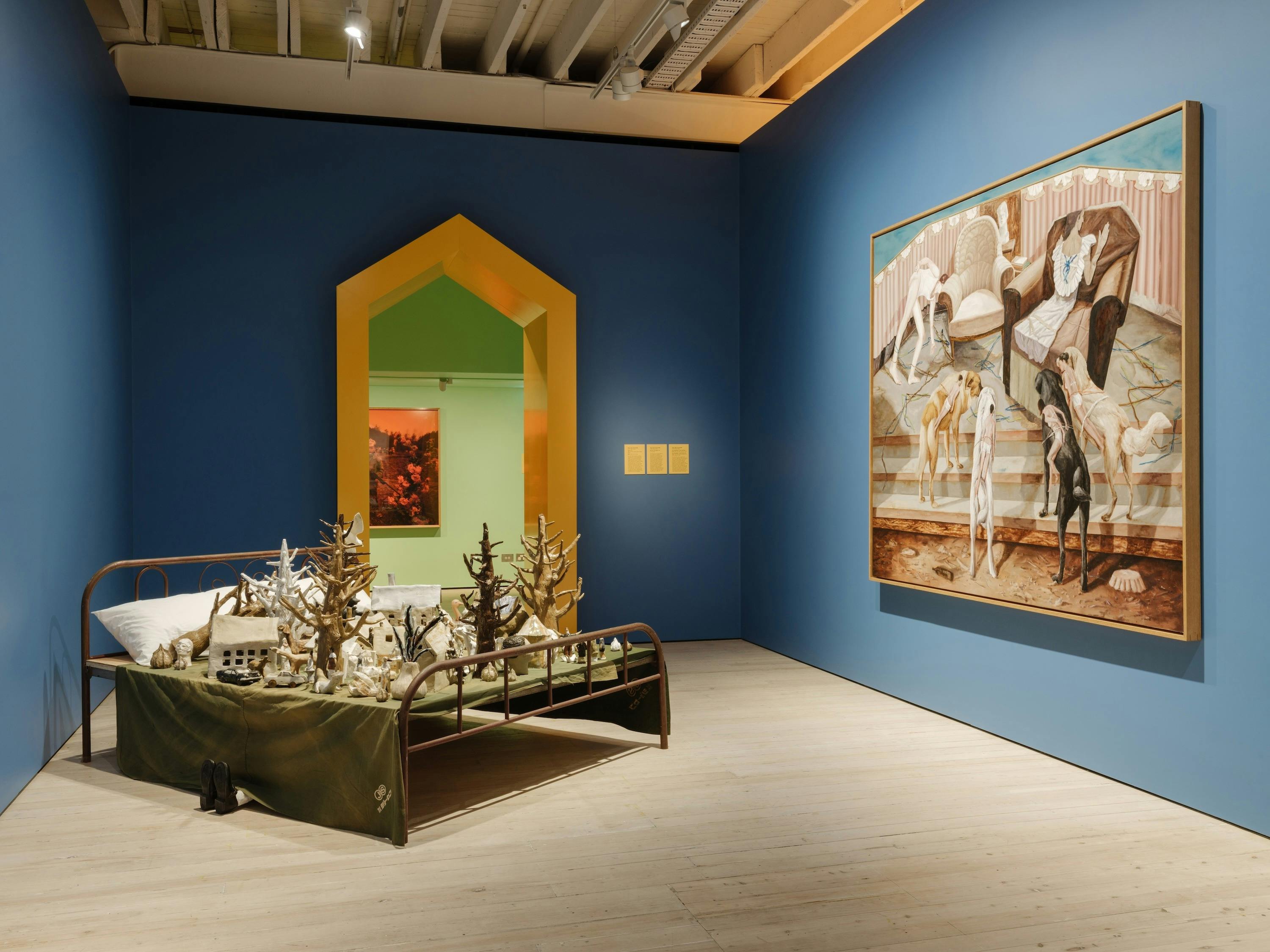 Blue walled gallery room with a sculpture of a bed and a canvas on the wall.