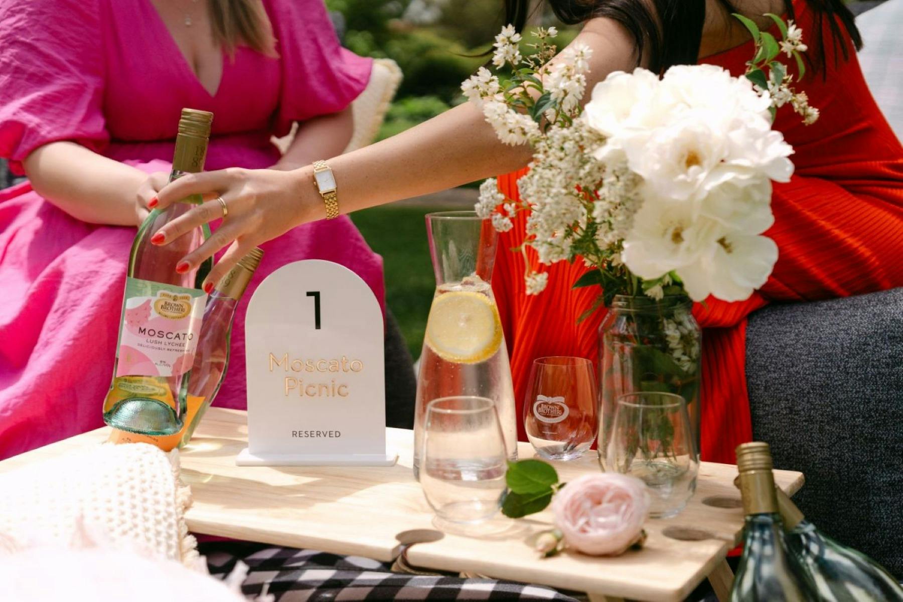 two people holding a wine bottle with glasses on a picnic table with a vase with white flowers