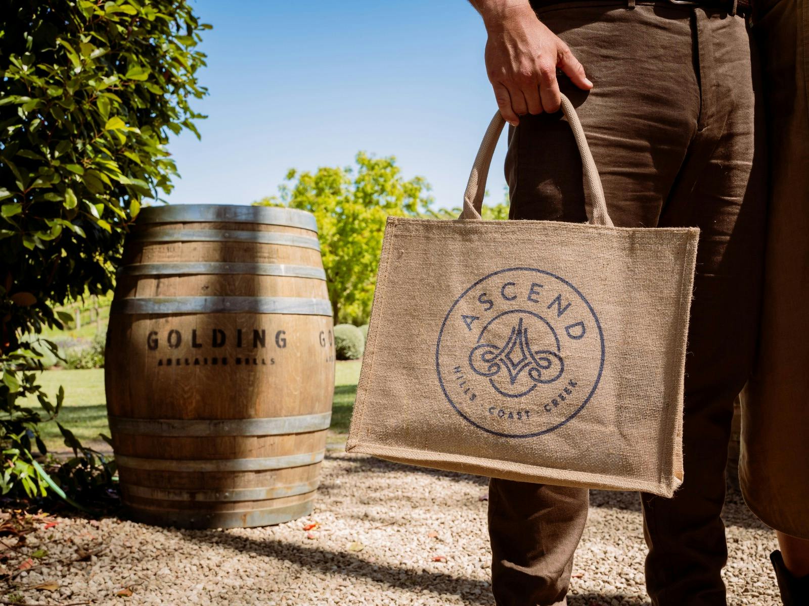 A close up image of a man holding an Ascend branded, hessian, tote gift bag, in a garden setting.