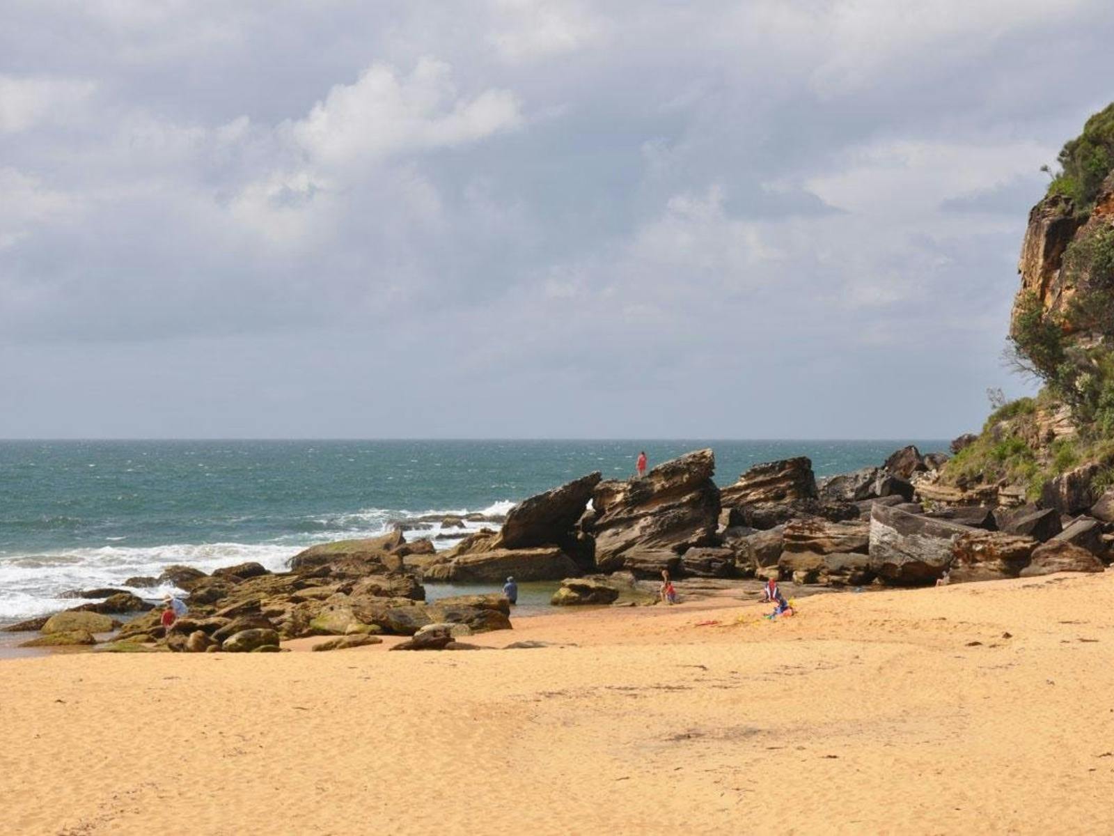 People swimming and standing on rocks beside a sandy beach and coastal cliff.