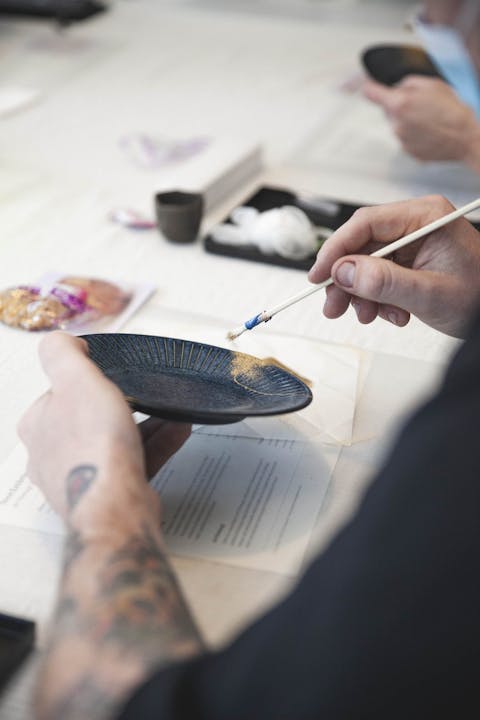 Picture shows hand holding a black dish with the other hand holding a brush with gold