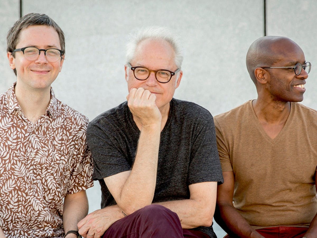 Three males sitting side by side. Two are smiling at the camera and one is looking away.