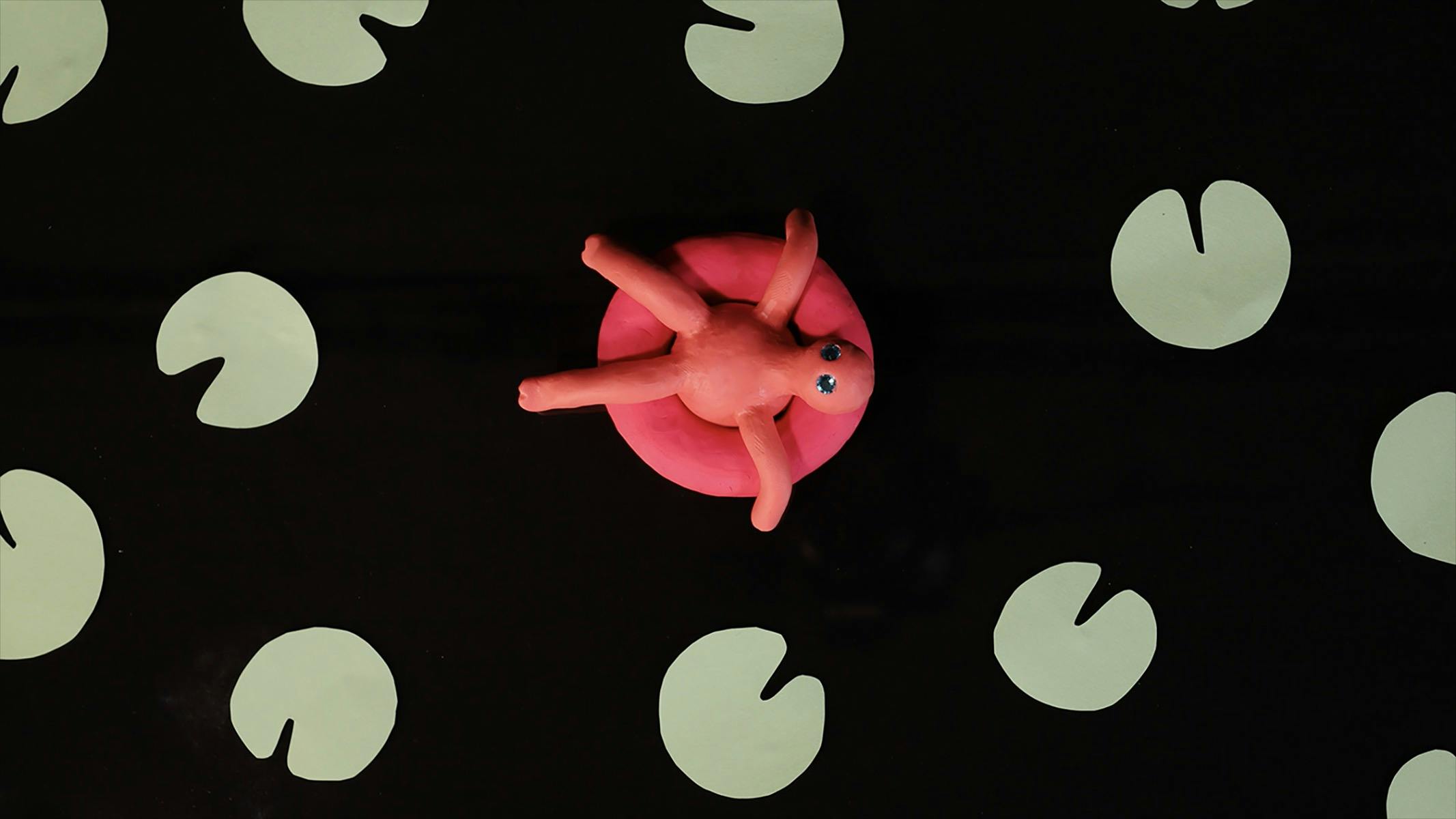 A video still image of a pink clay character sitting in a pool float surrounded by green lily-ads
