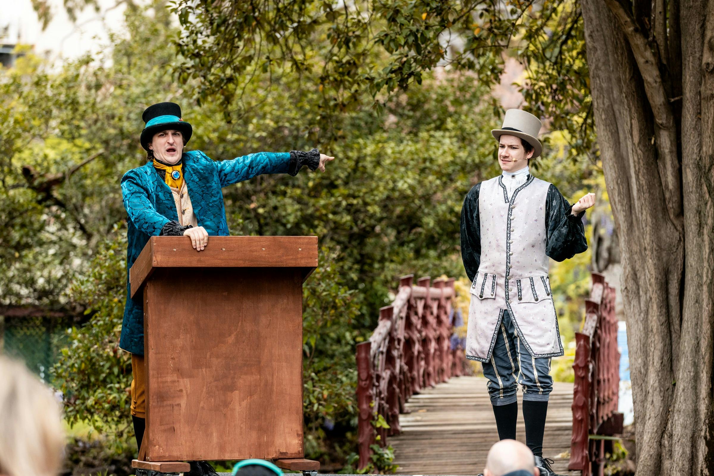 A man in a green coat points accusatorially at another man dressed in an elaborate costume.