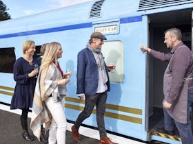 4 white adults walking next to a train carrying glasses, a waiter welcomes then onboard the train