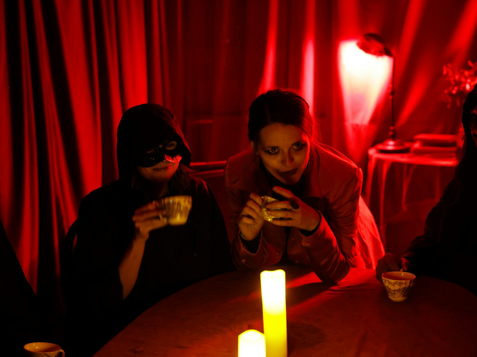 A person in a black cloak and mask, and a person in a red jacket holding teacups in a dim red room