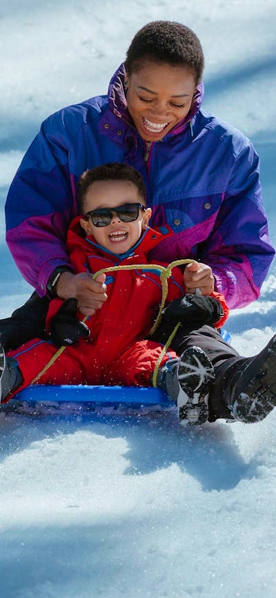A mother and son riding a toboggan down a slope covered in snow, with big smiles on their faces.