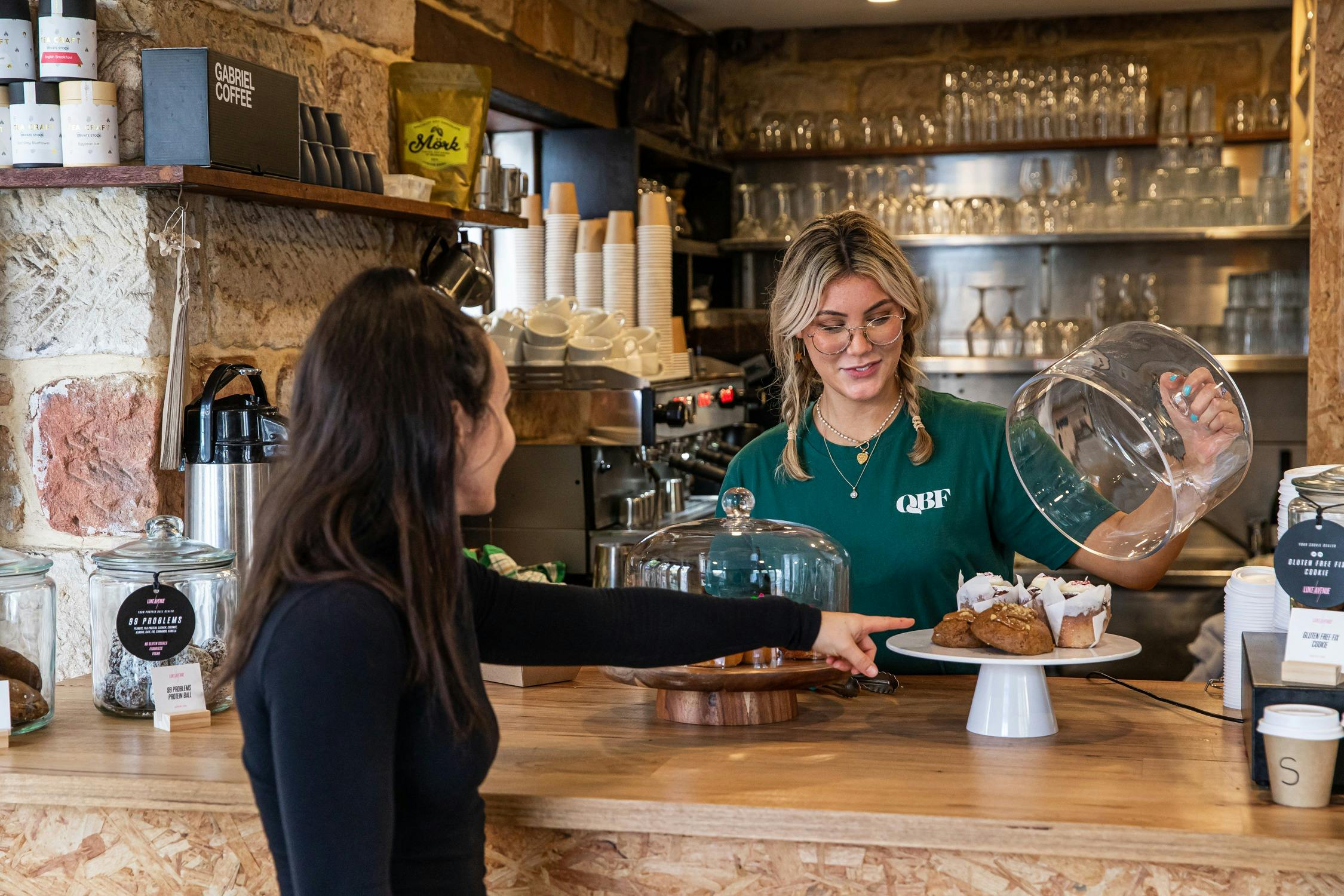Takeaway counter with staff member opening a cloche of pastries and a customer pointing at a cake