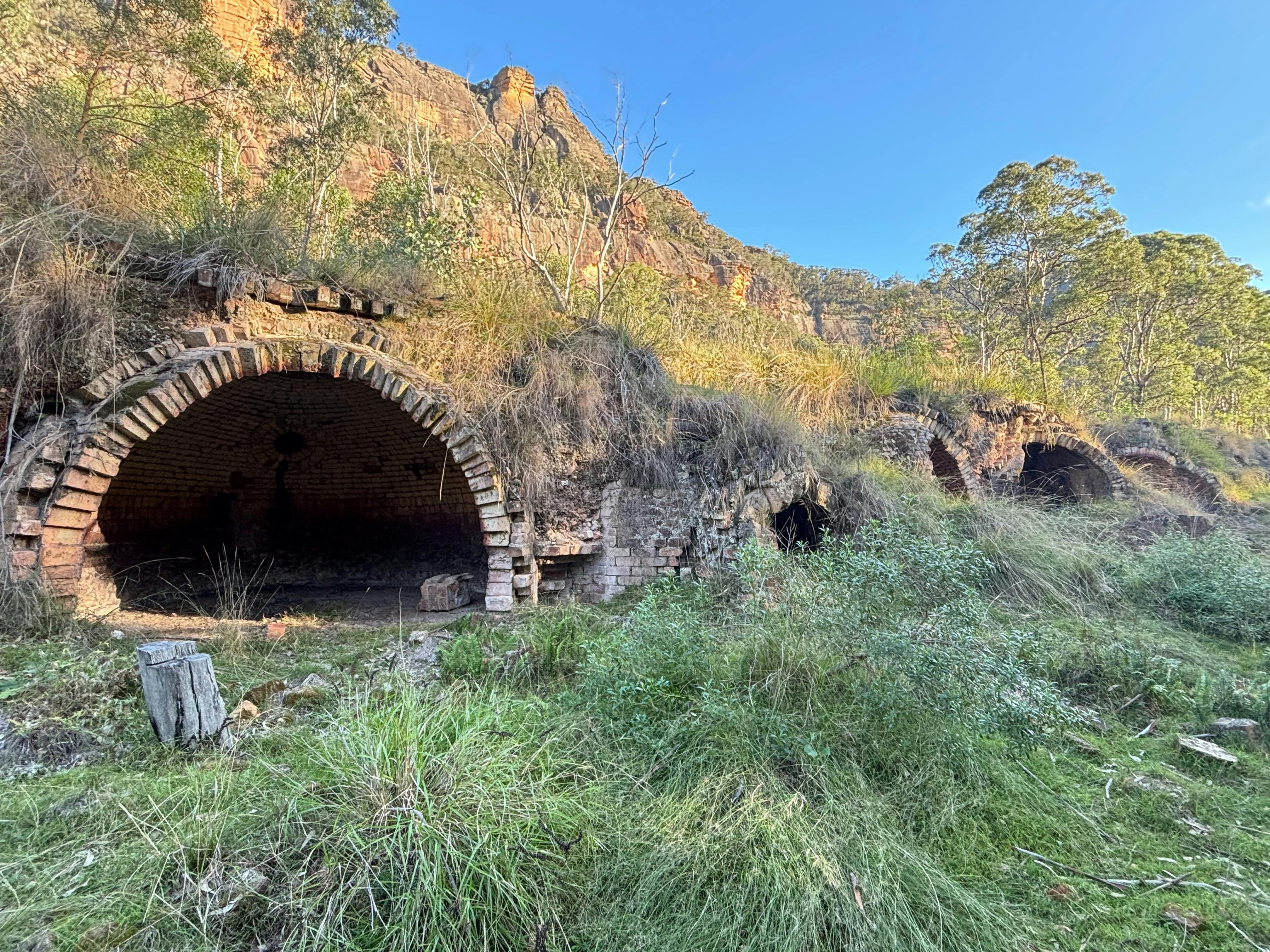 A row of large brick ovens covered in grass and natural bush with high red stand stone cliffs behind