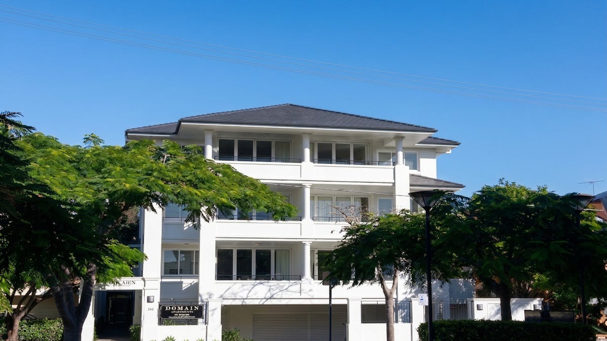 A white three-storey apartment building with balconies on each level, framed by mature trees, with a