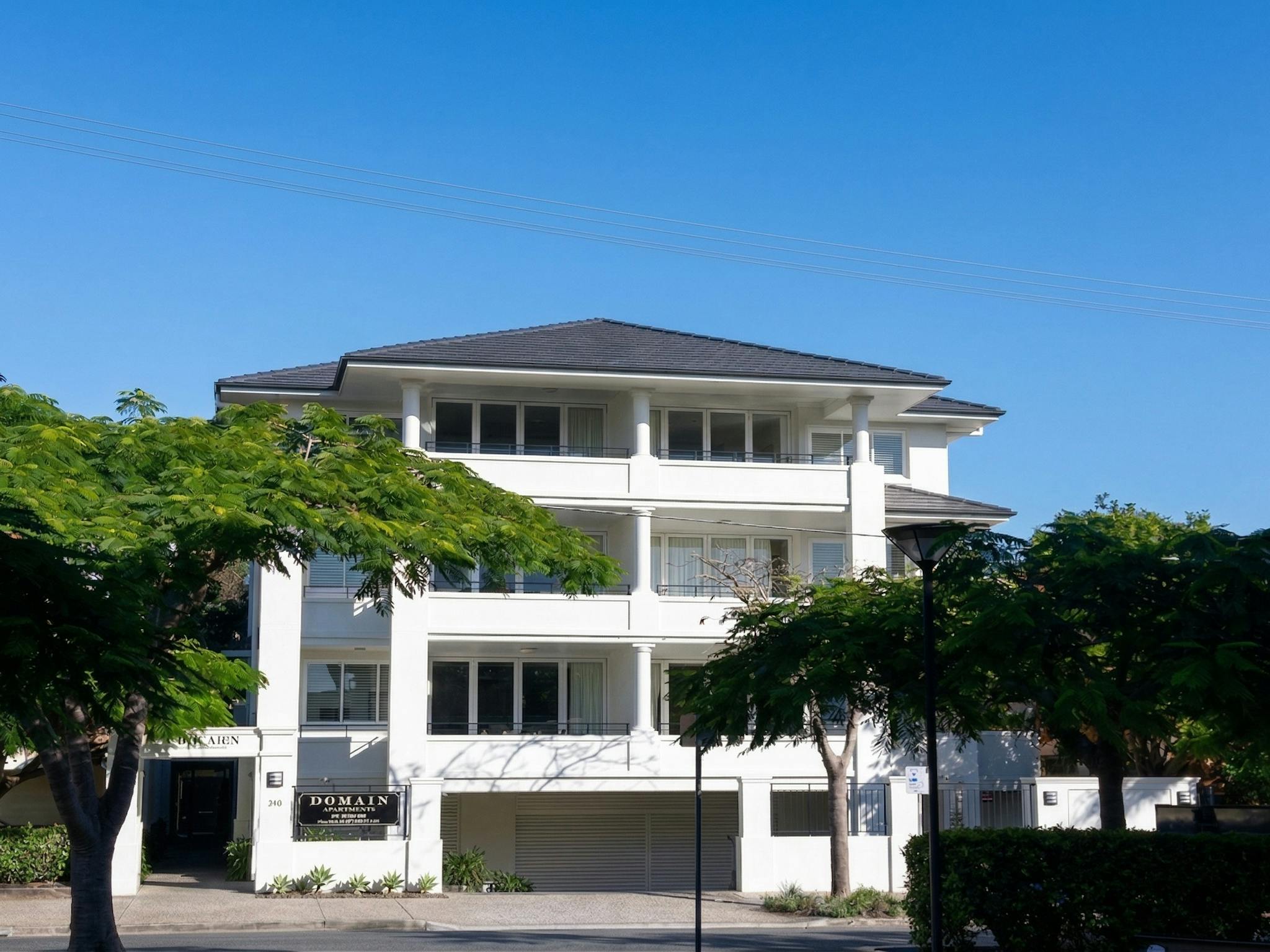 A white three-storey apartment building with balconies on each level, framed by mature trees, with a A white three-storey apartment building with balconies on each level, framed by mature trees, with a