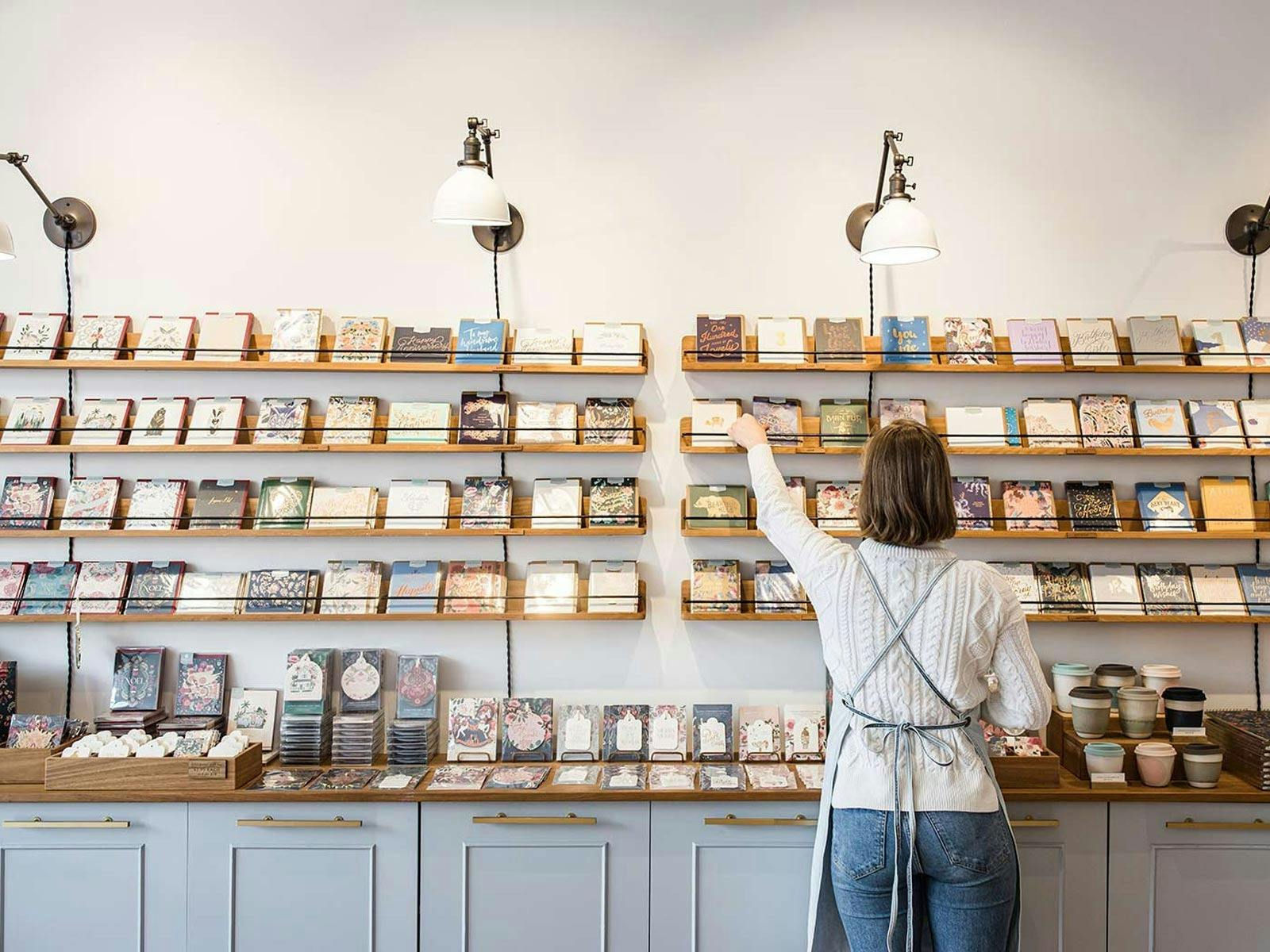 Woman arranging greeting cards on shelves in a beautifully styled stationery store interior.