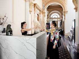 A woman in black at a reception desk hands a ticket to another woman in a Museum foyer with statues