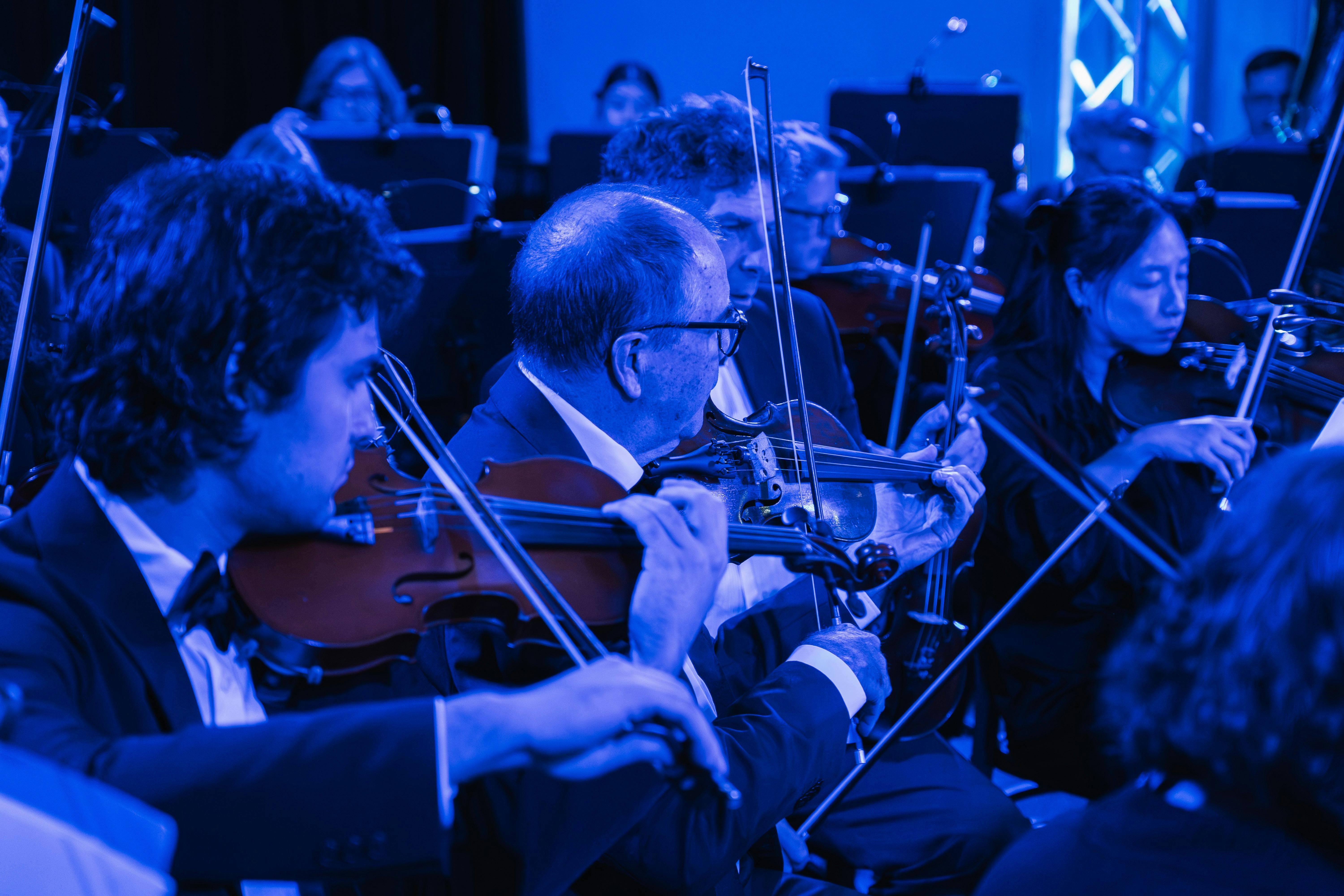 Orchestra members in blue light play violins, dressed in tuxedos