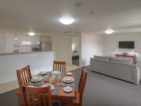 Four seater dining table in foreground. Lounge with TV on right and full kitchen in background.