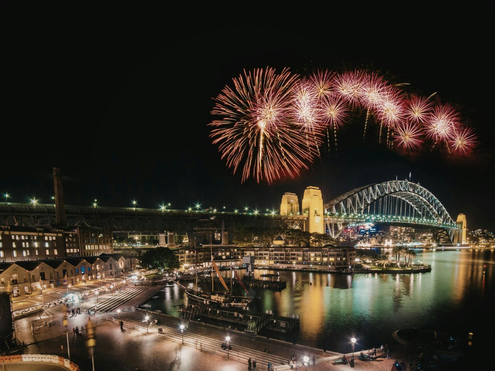 Fireworks bursting over Sydney Harbour Bridge at night, The Rocks precinct below