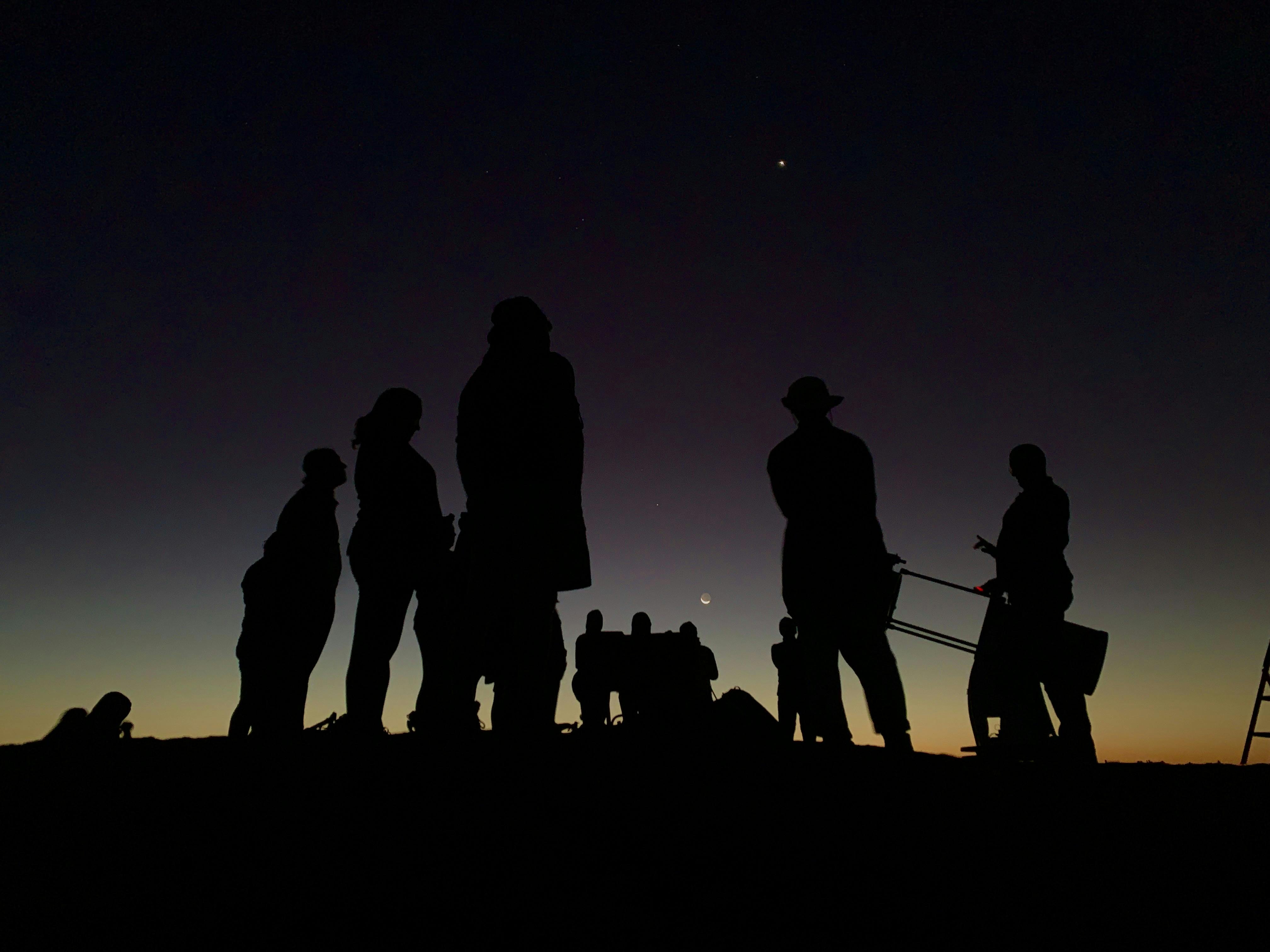 Silhouette image of hikers at dusk looking up at stars, sky is deep purple and yellow hues