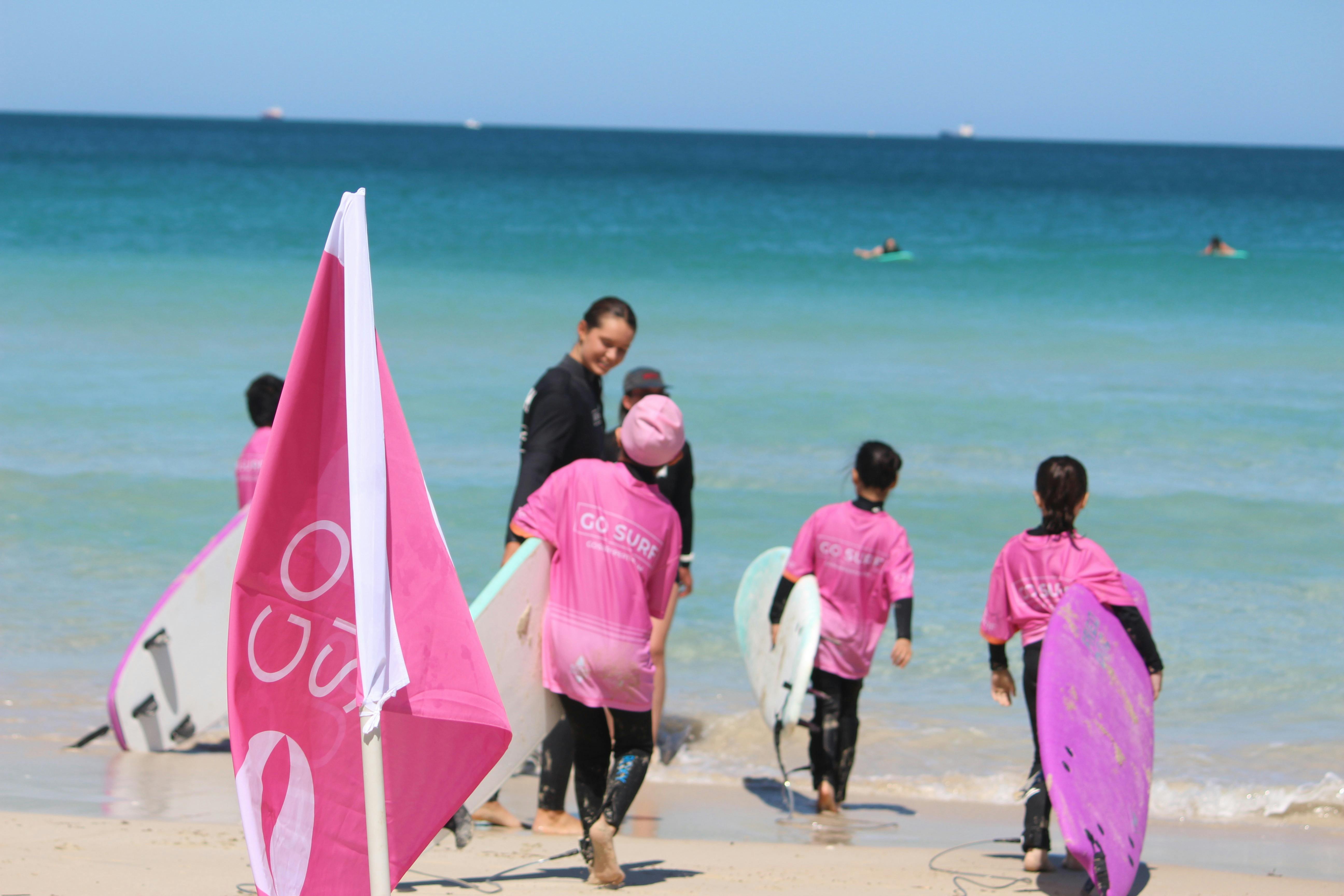 Young girls carrying surfboards follow their female surf instructor into the water on a sunny day