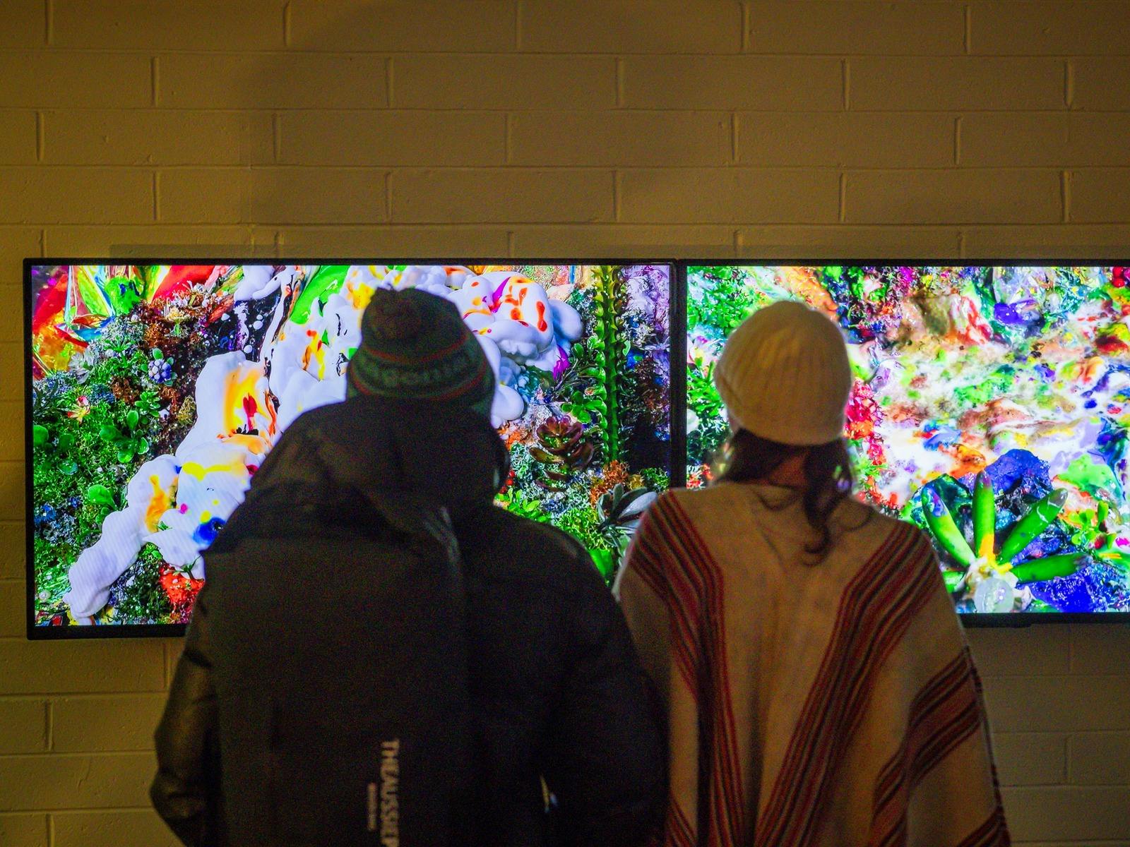 Two people stand infront of an screen based colourful video artwork by Jacob Leary.