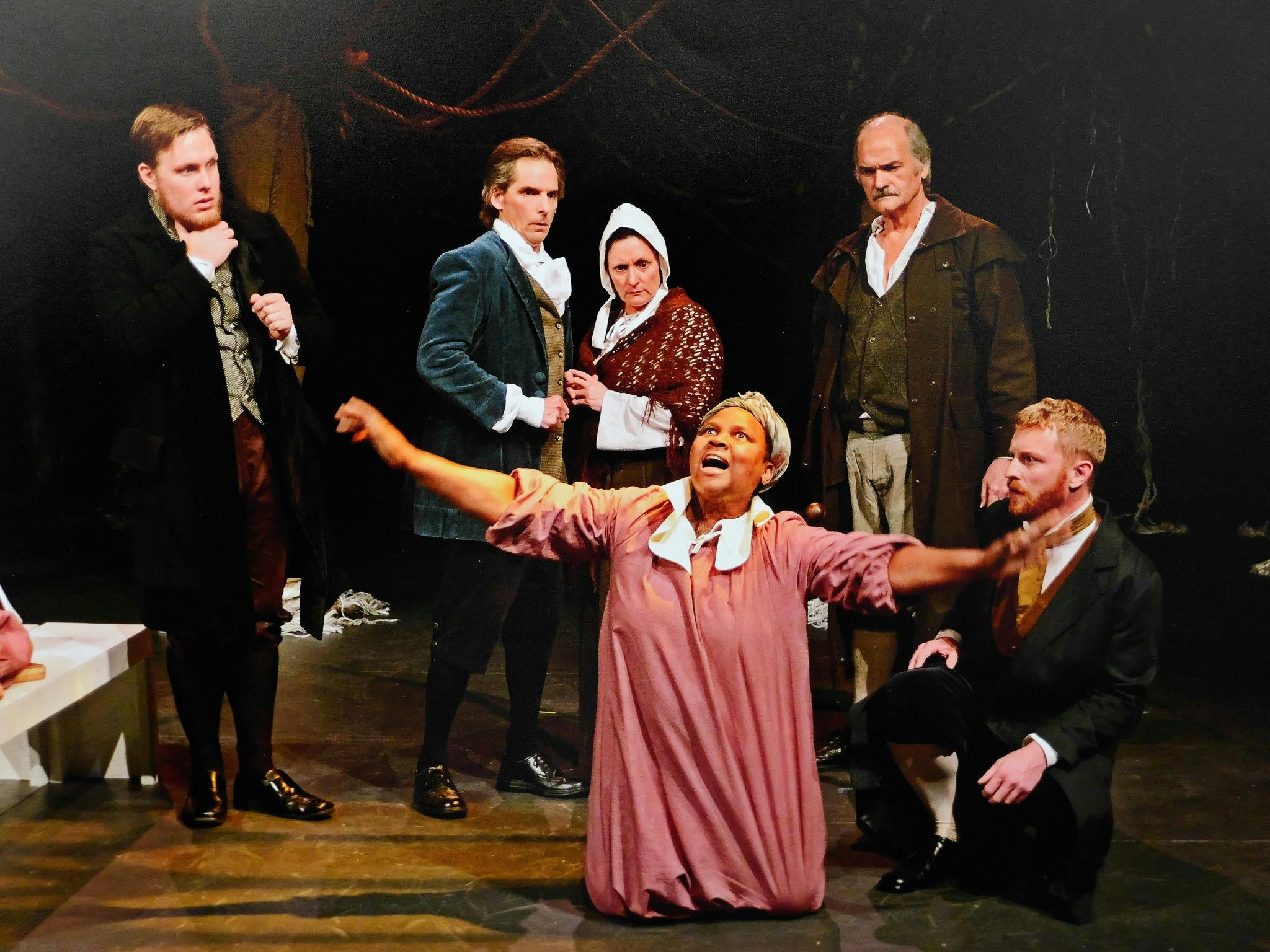 a young woman in pink dress is on her knees as 5 others in 1690s period costume look on in fear