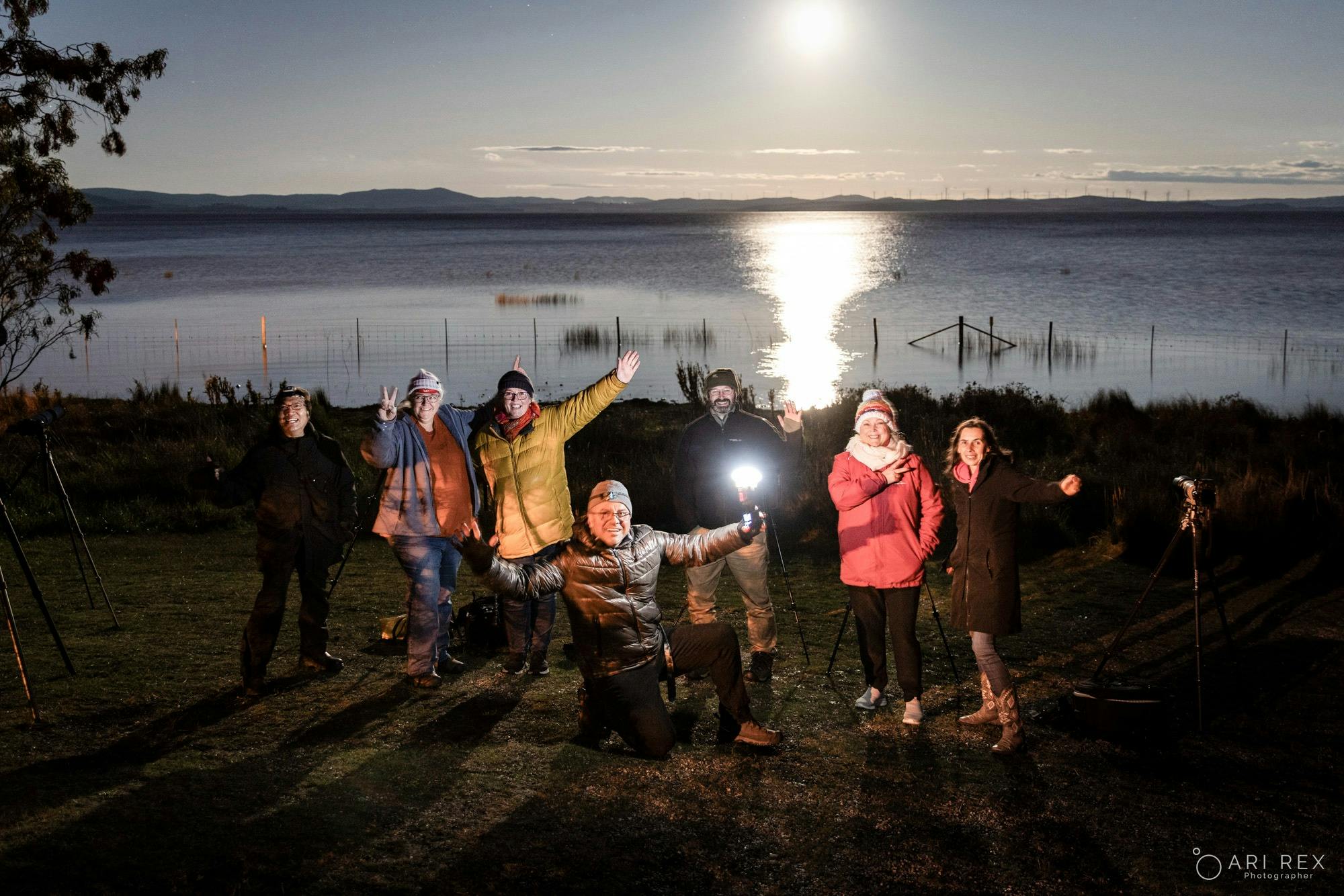 Photographers pose beside a lake under the full moon, with tripods and a camera flash.