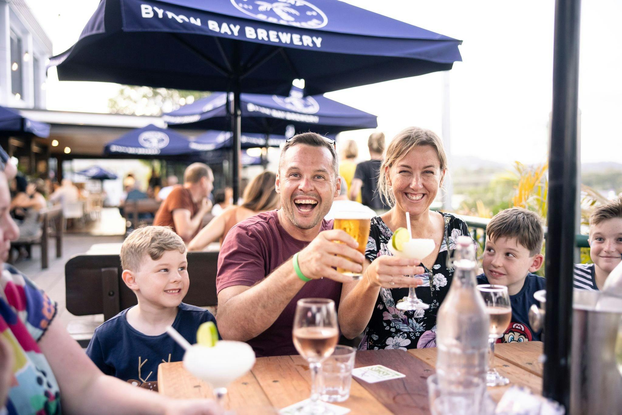 Mum, Dad and two kids sitting at an outdoor table smiling at the camera with drinks raised