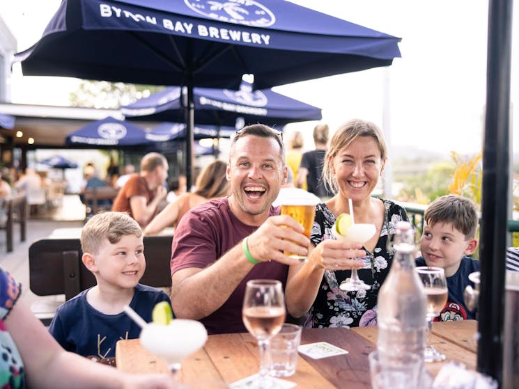 Mum, Dad and two kids sitting at an outdoor table smiling at the camera with drinks raised