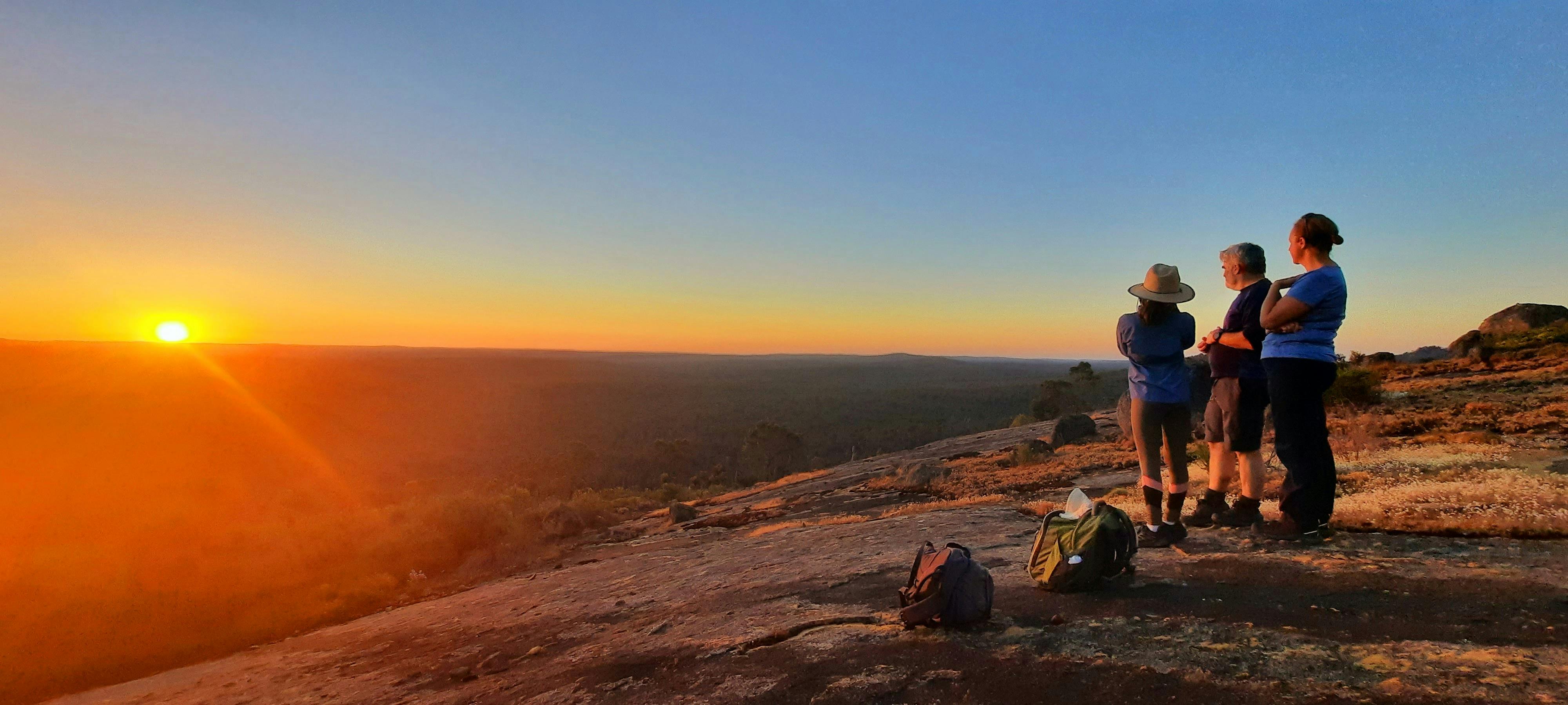 3 hikers stand on top of hill admiring beautiful orange sunset, sky is a clear warm blue