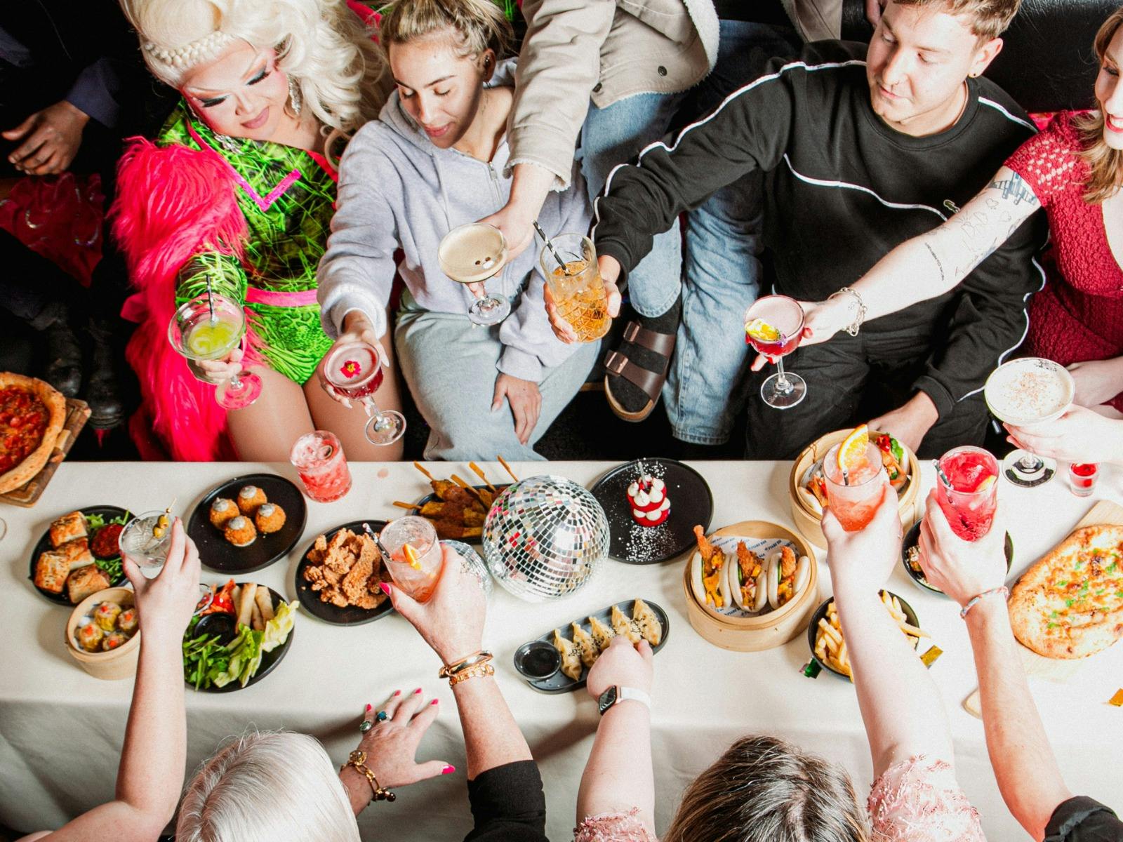 Guests raise cocktails around a table of shared food with a drag performer.