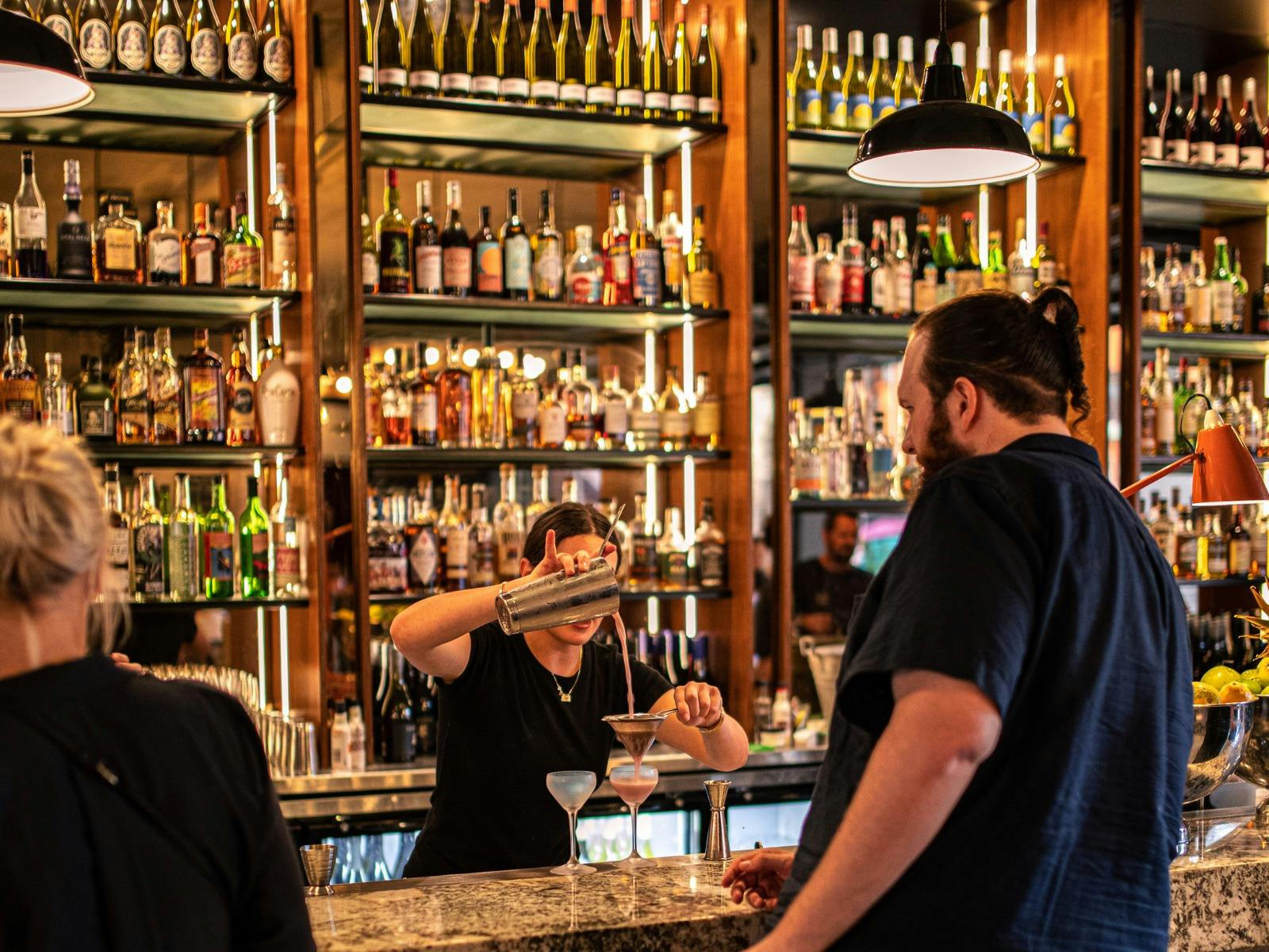 Bartender pouring cocktail into glass at bar with people waiting at bar