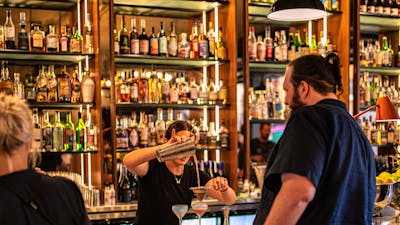 Bartender pouring cocktail into glass at bar with people waiting at bar