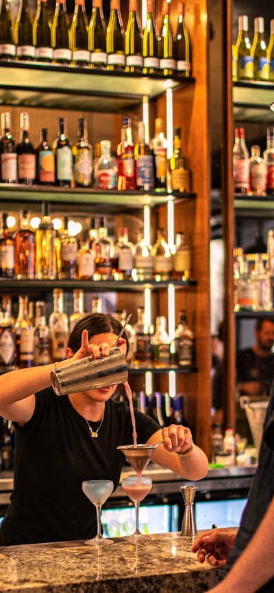 Bartender pouring cocktail into glass at bar with people waiting at bar