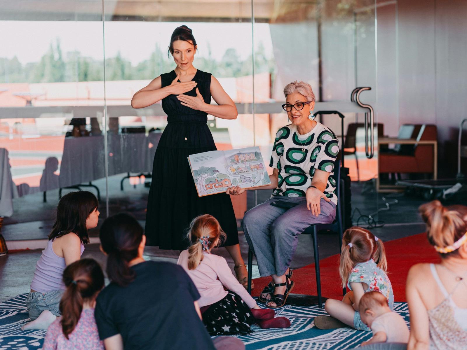 An Auslan interpreter standing next to a person reading a book to a group of children sitting down