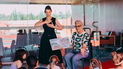 An Auslan interpreter standing next to a person reading a book to a group of children sitting down