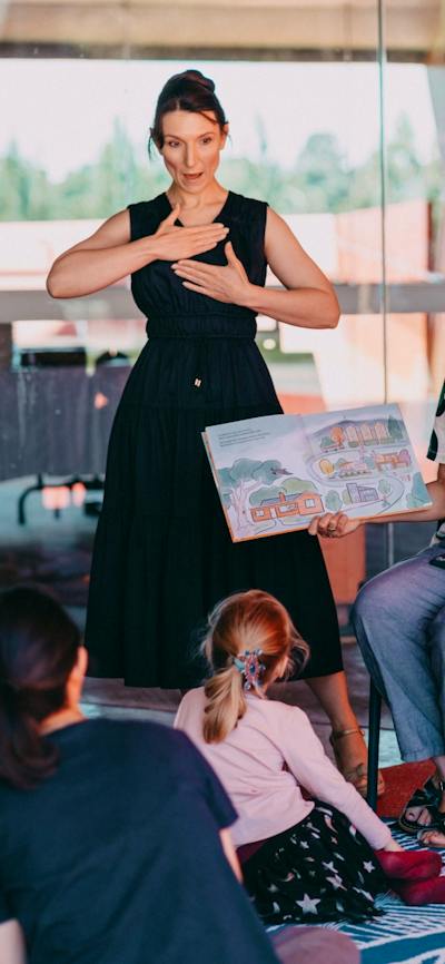An Auslan interpreter standing next to a person reading a book to a group of children sitting down