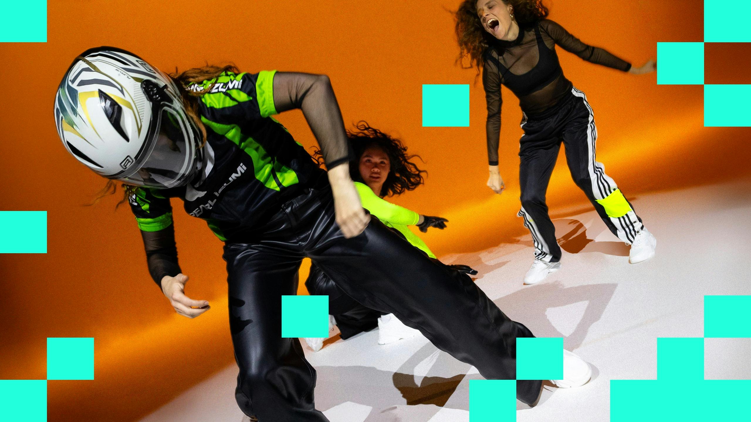 Three dancers  wearing V8 racing costumes, one in a helmet, in front of an orange background.